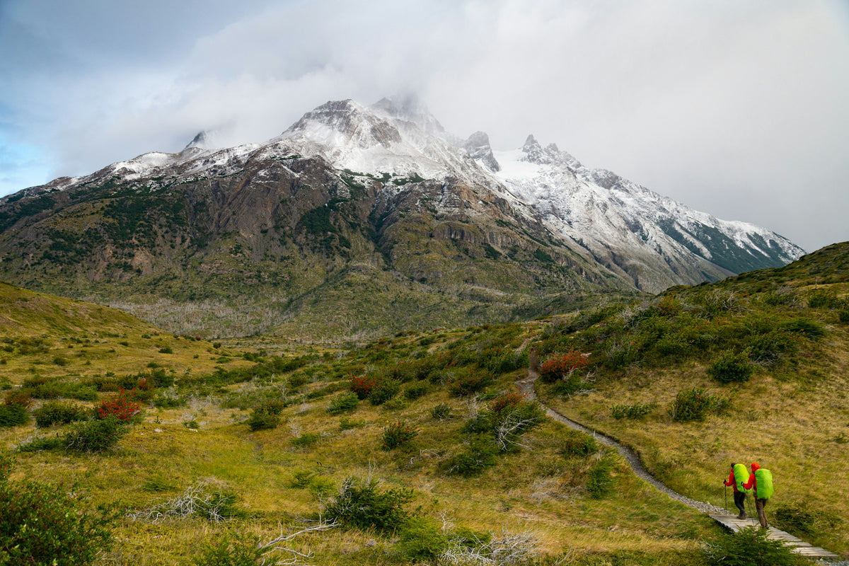 Two trekkers with backpacks hiking toward the snow-capped peaks of Torres del Paine, Chile
